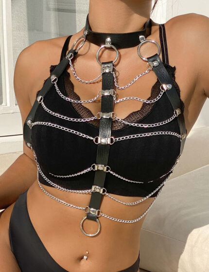 Black faux leather body harness with silver chains and metal O-rings worn over a black lace bralette by a model sitting in sunlight